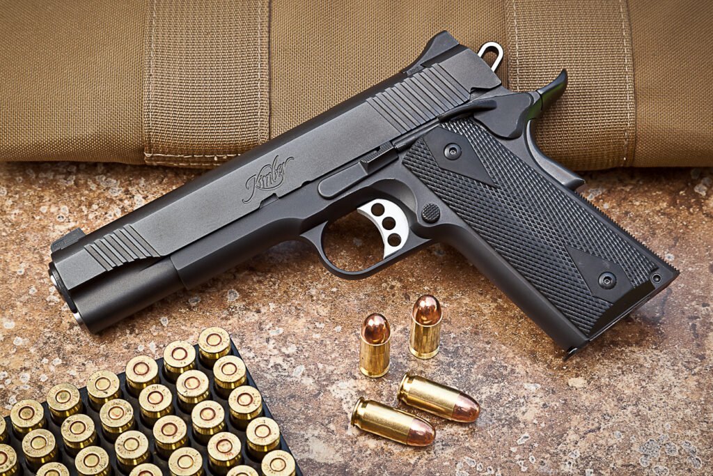 Kimber 1911 handgun with black textured grips displayed alongside .45 ACP ammunition and a tactical case in the background.