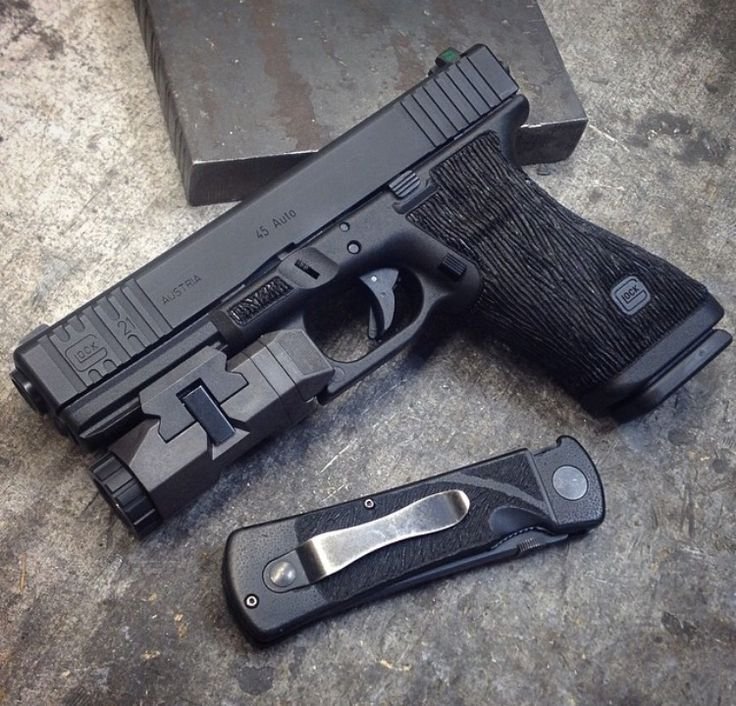 A black semi-automatic pistol with a mounted flashlight and a textured grip, placed on a rough surface next to a folding knife.