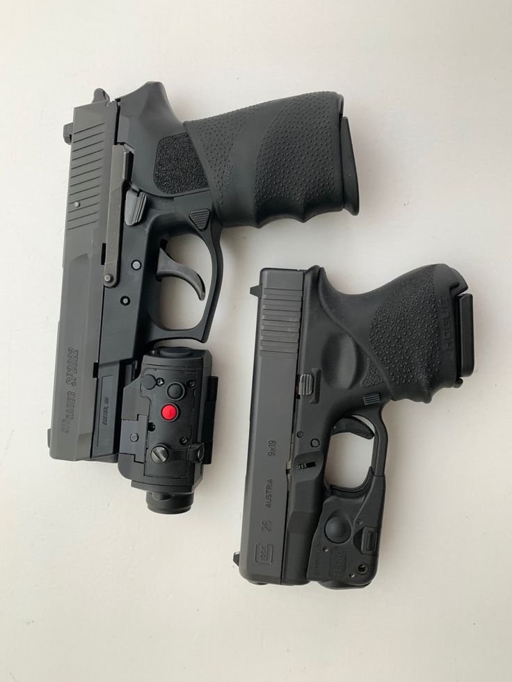 Two black semi-automatic handguns placed side by side on a light surface. The handgun on the left features a mounted tactical light or laser attachment.